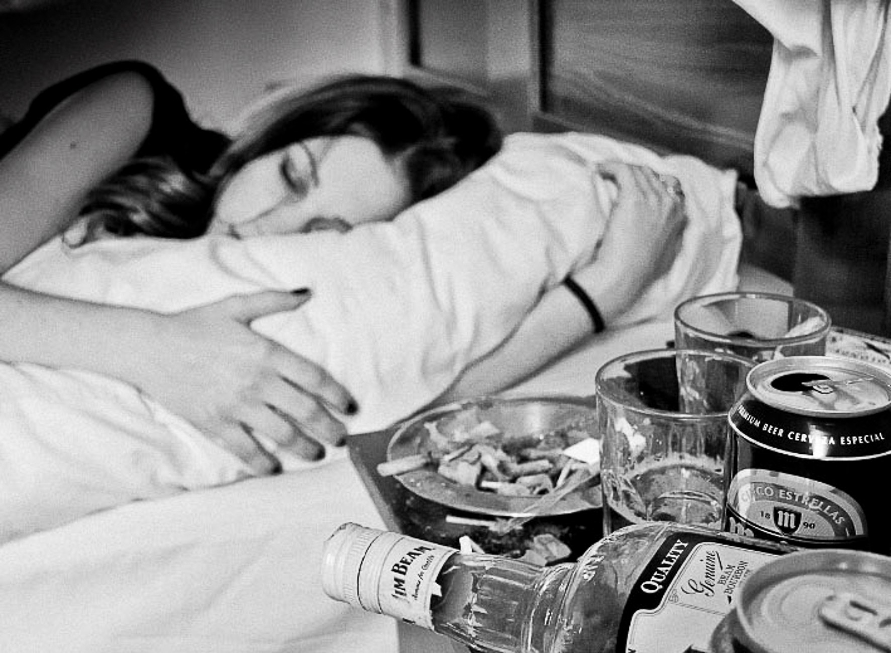 A woman asleep hugging a pillow, with an ashtray, glasses and cans on the bedside table.