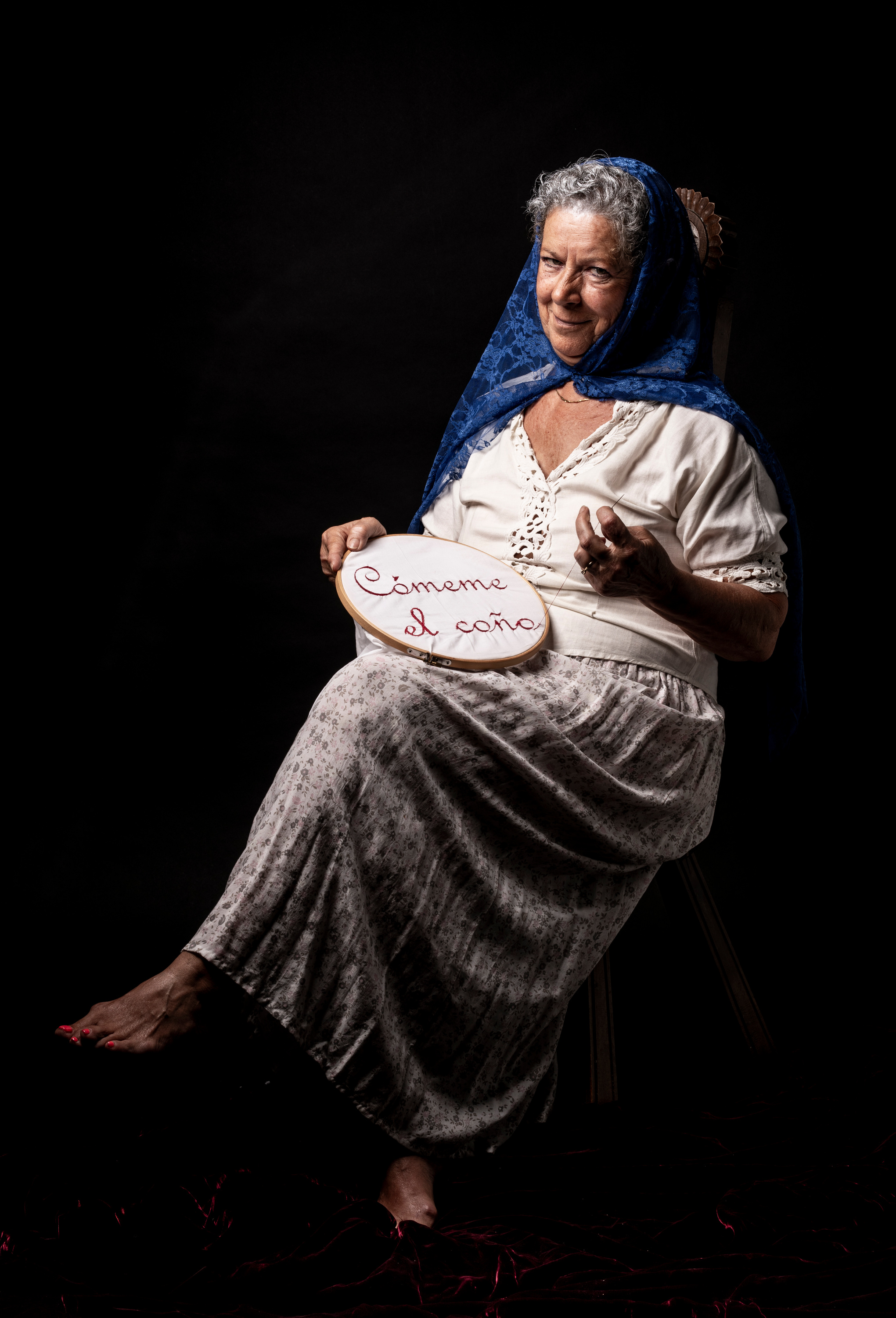 "La abuela". An older woman in a blue headscarf, seated, embroidering the phrase "Cómeme el coño" — "Lick my cunt" — on a hoop.