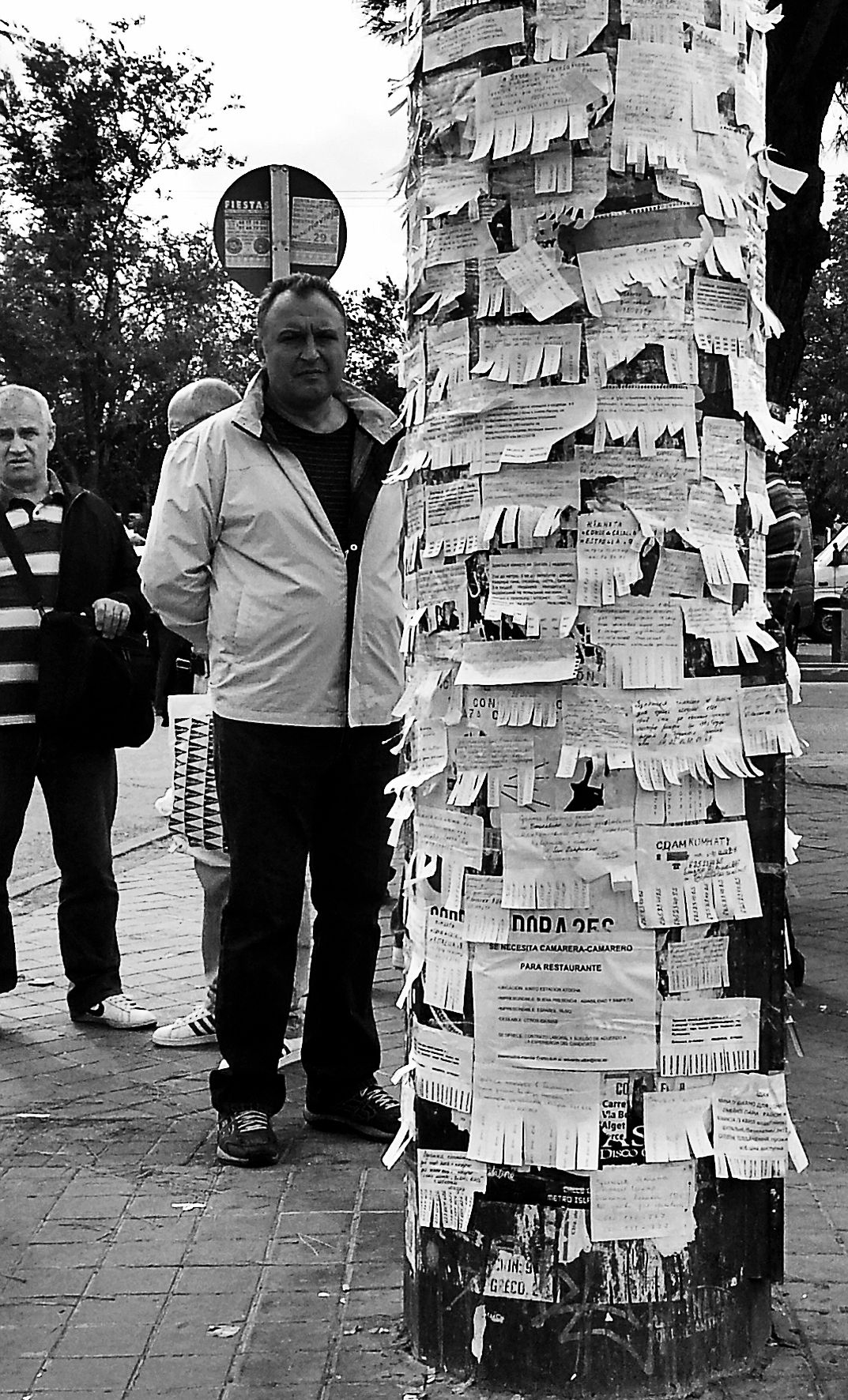 A man stands beside a street post plastered with paper ads and their tear-off tabs. Black and white photograph by Javier Jimeno.